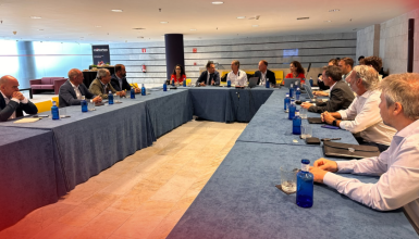Group of professionals seated around a U-shaped blue-clothed conference table in a modern meeting room, engaged in discussion.