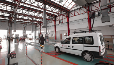 Industrial vehicle maintenance bay with a white service van and a technician walking toward the back, red structural columns and skylights overhead.