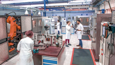 Industrial lab scene: technicians in white coats work with large automated machinery and control panels; a robotic arm and various tools are arranged on a workbench, while two scientists discuss a component in the center of the room; the space is tidy with tiled floors and safety mats along the walkways.