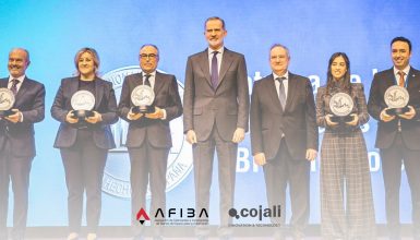 Six professionals in formal attire stand on a stage, each holding a round award plaque, with a blue backdrop and logos at the bottom.