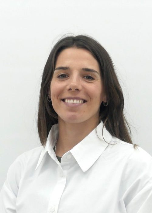 Smiling woman in a white button-down shirt stands with hands clasped in front against a pale gray background.