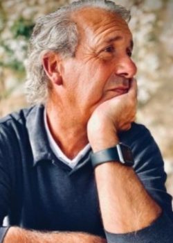 Older man with gray hair sits outdoors, resting his chin on his hand and gazing thoughtfully to the side.