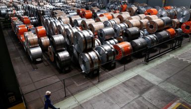 Industrial warehouse with stacked steel coils in orange, silver, and gray; a worker in a hard hat walks along a railing.