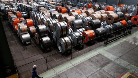 Industrial warehouse with stacked steel coils in orange, silver, and gray; a worker in a hard hat walks along a railing.